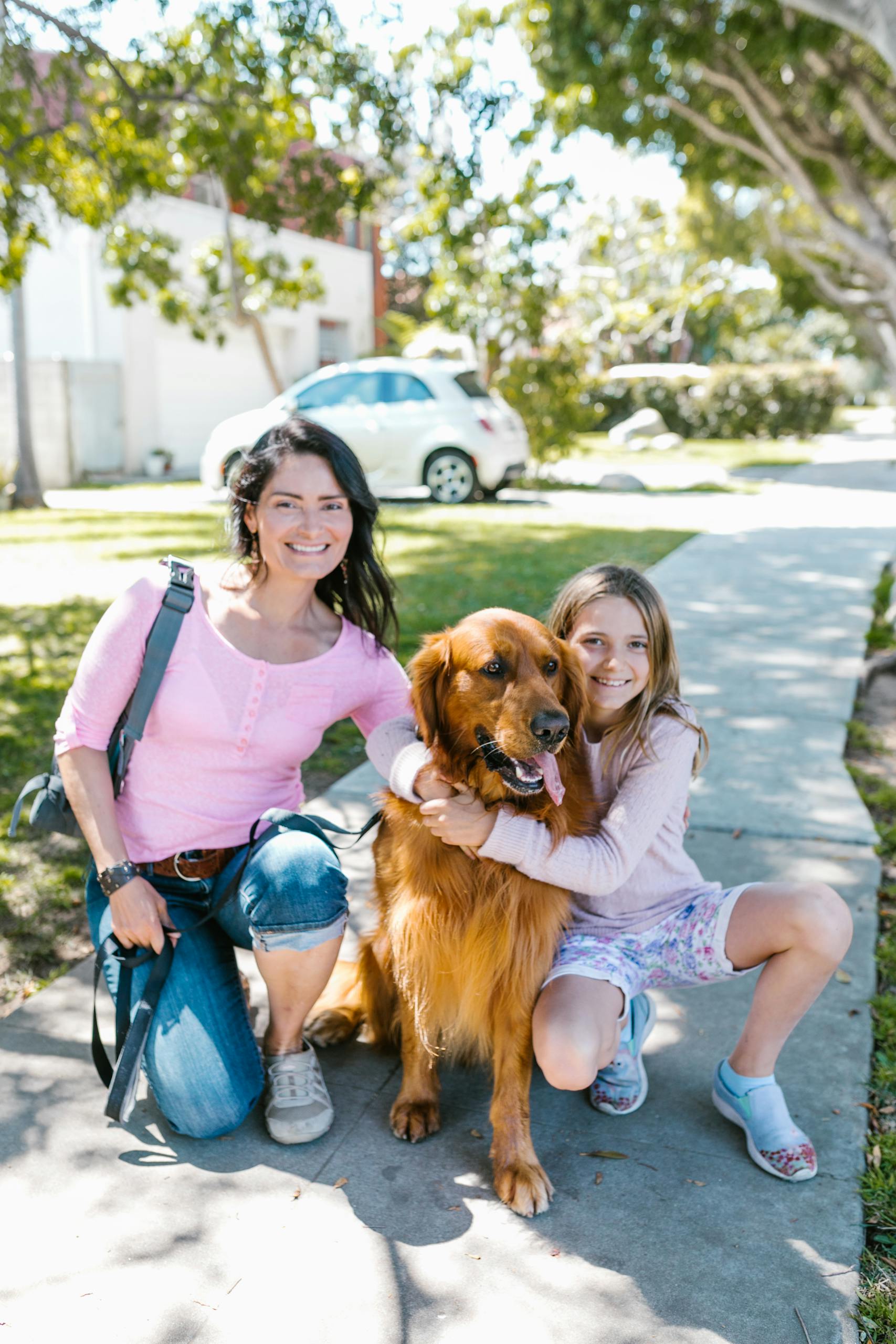 A woman and girl smile with their golden retriever dog outdoors on a sunny day.